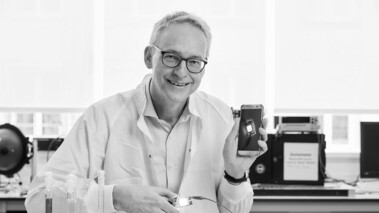 Älterer Wissenschaftler im weißen Kittel sitzt in einem modernen Labor und lächelt in die Kamera.